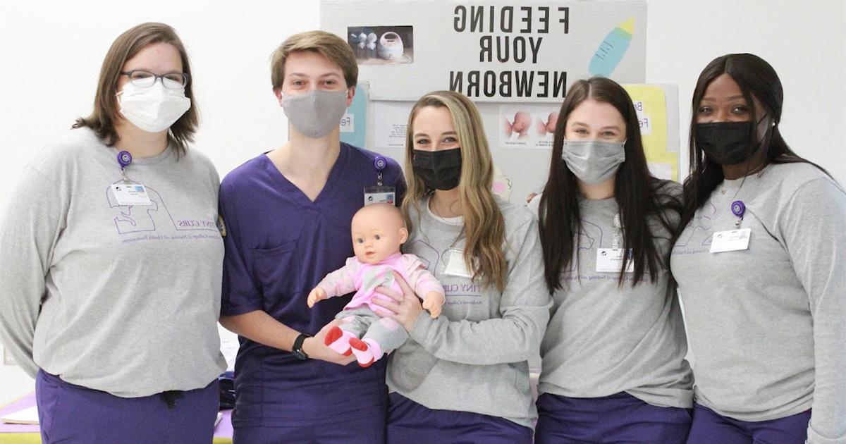 Nursing students stand in front of a posterboard explaining how to feed your newborn. They are all wearing masks, purple scrubs, and grey shirts.